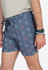 Bañador en azul marino con un patrón de calaveras y huesos cruzados en azul claro y rojo. Cuenta con una cintura con cordón ajustable y un pequeño parche con el logo en la pierna.