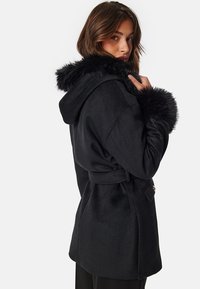 Black coat with a fur-lined hood, belted waist, and side pockets. Soft fabric with a smooth texture and minimalistic design details.