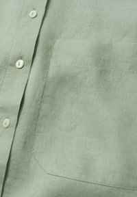Lichtgroen linnen overhemd met een knoopsluiting aan de voorkant, kleine ronde knopen en een borstzakje aan de linkerkant. Glad, gestructureerd stof.