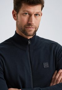 Navy zip-up sweater with a high neckline, featuring a textured pattern and a small logo patch on the left chest. Soft fabric and tailored fit.