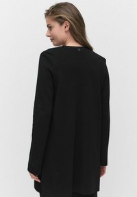 Kvinna med långt hår som bär en lång svart kappa, vänd något åt sidan, mot en enkel ljus bakgrund.