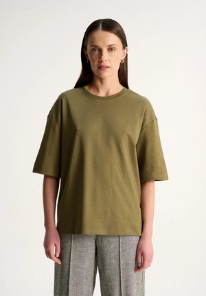 Kobieta z prostymi ciemnymi włosami ma na sobie za dużą oliwkowozieloną koszulę z krótkim rękawem i szare plisowane spodnie, stoi na tle jednolitego jasnego tła.