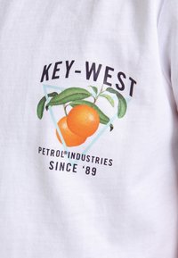 Bílý bavlněný tričko s grafikou dvou pomerančů, zelenými listy a textem: "KEY-WEST PETROL INDUSTRIES SINCE '89" pod nimi.