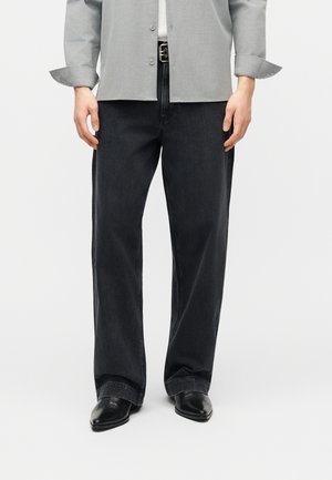 Άνδρας που φοράει φαρδιά μαύρα τζιν, μαύρα μυτερά παπούτσια, ανοιχτό γκρι πουκάμισο με κουμπιά και μανίκια γυρισμένα, στέκεται μπροστά σε λευκό φόντο.