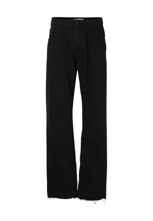 Homme portant un jean noir large et des chaussures noires, debout devant un fond uni, les mains relâchées le long du corps.