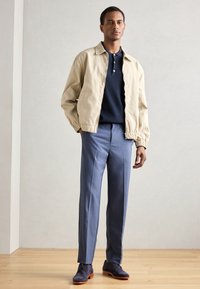 Giubbotto leggero beige con polsini elastici, polo blu navy, pantaloni leggeri su misura blu chiaro e scarpe blu scuro con suole chiare.