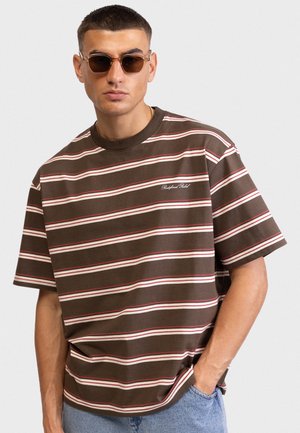 Καφέ ριγέ μπλούζα με λευκές και κόκκινες γραμμές, κοντά μανίκια, ριμπ λαιμόκοψη και κεντημένο λογότυπο της μάρκας στο στήθος. Φορεμένη με γυαλιά ηλίου.