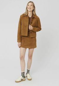 Veste en daim marron avec deux poches avant et des boutons, assortie d'une jupe marron. Portée avec des chaussettes à motifs léopard et des baskets beiges.