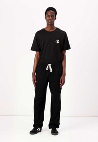 T-shirt noir en coton avec un petit graphique sur la poitrine, associé à un pantalon de jogging noir avec cordon de serrage. Le mannequin porte des baskets noires.