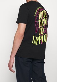 Černé bavlněné tričko s velkým žlutým a růžovým grafickým designem a textem „TVŮJ ČAS PODPOŘIT“ na zádech.