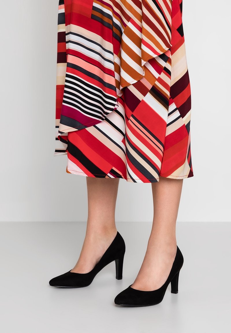 Jambes vêtues de talons hauts en daim noir, associées à une jupe sous le genou avec des rayures diagonales vibrantes rouges, orange, roses et noires.
