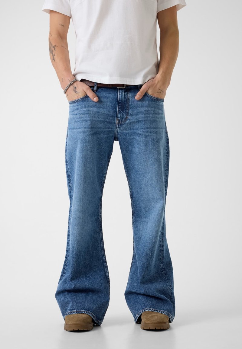 Mężczyzna w jasnoniebieskich szerokich dżinsach, brązowych butach, białej koszulce, z rękami w kieszeniach, stojący na jednolitym białym tle.