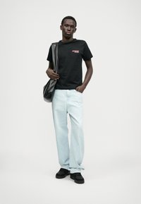 Svart bomulls t-skjorte med rødt grafisk trykk, lyseblå denimjeans og svarte sko. Modellen holder en svart veske og står foran en ensfarget bakgrunn.