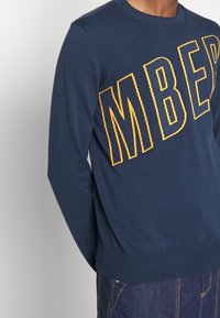 Jersey de color azul marino con grandes letras amarillas "MBER." Presenta un cuello redondo, puños y dobladillo acanalados. Está hecho de una tela suave y elástica.
