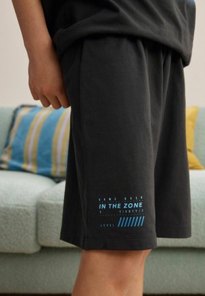 Person iført sorte shorts med blå tekst, der lyder "Game over In the Zone Level", stående indendørs nær en sofa med stribede puder.