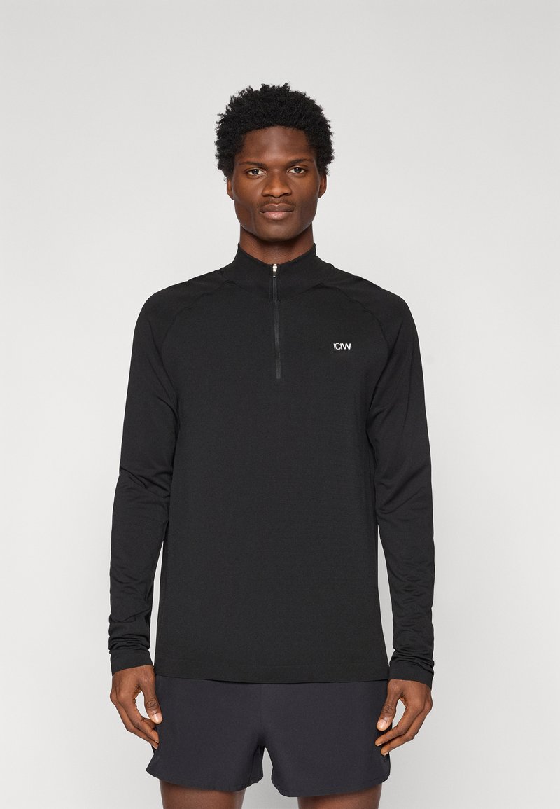 Μαύρο αθλητικό τοπ με μακριά μανίκια, γιακά με μισό φερμουάρ και ανακλαστικό λογότυπο, με λεία υφή και εφαρμοστή σχεδίαση.