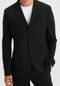 Svart blazer i slät tyg med slagkrage, tre knappar, två patchfickor och en smal passform. Bärs över en vit t-shirt.