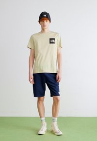 T-shirt in cotone beige con patch logo nero, pantaloni corti blu navy, sneakers bianche e un berretto di lana nero. Outfit semplice e casual su uno sfondo chiaro.