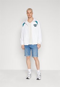 Chaqueta ligera blanca con acentos en verde azulado, camiseta gris debajo, pantalones cortos de mezclilla y zapatillas grises, de pie contra un fondo neutral.