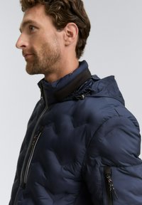 Profil av en man med kort, krulligt hår iförd en mörkblå vadderad jacka med dragkedjefickor och justerbar huva.