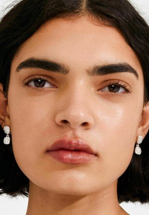 Pendientes colgantes de plata con una superficie texturizada, que presentan dos elementos ovalados interconectados. Usados por una persona de cabello oscuro.