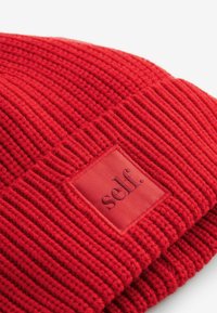 Rød strikket hue med ribbet tekstur, prydet med et rektangulært stofmærke prægede med "self." på fronten til branding.