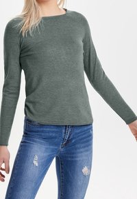 Grünes Langarmoberteil in lässigem Schnitt mit rundem Halsausschnitt. Kombiniert mit blauen, verwaschenen Jeans, die eine entspannte Textur zeigen.