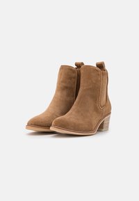 Braune Wildleder Stiefeletten mit runder Zehenform, seitlichen elastischen Einsätzen und einem gestapelten Absatz. Sie verfügen über eine glatte Oberfläche und eine dezente Textur.