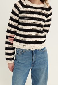 Gehaakt sweater met zwarte en crèmekleurige strepen. Heeft een open brei ontwerp en een gegolfde zoom. Gecombineerd met lichtblauwe denim jeans.