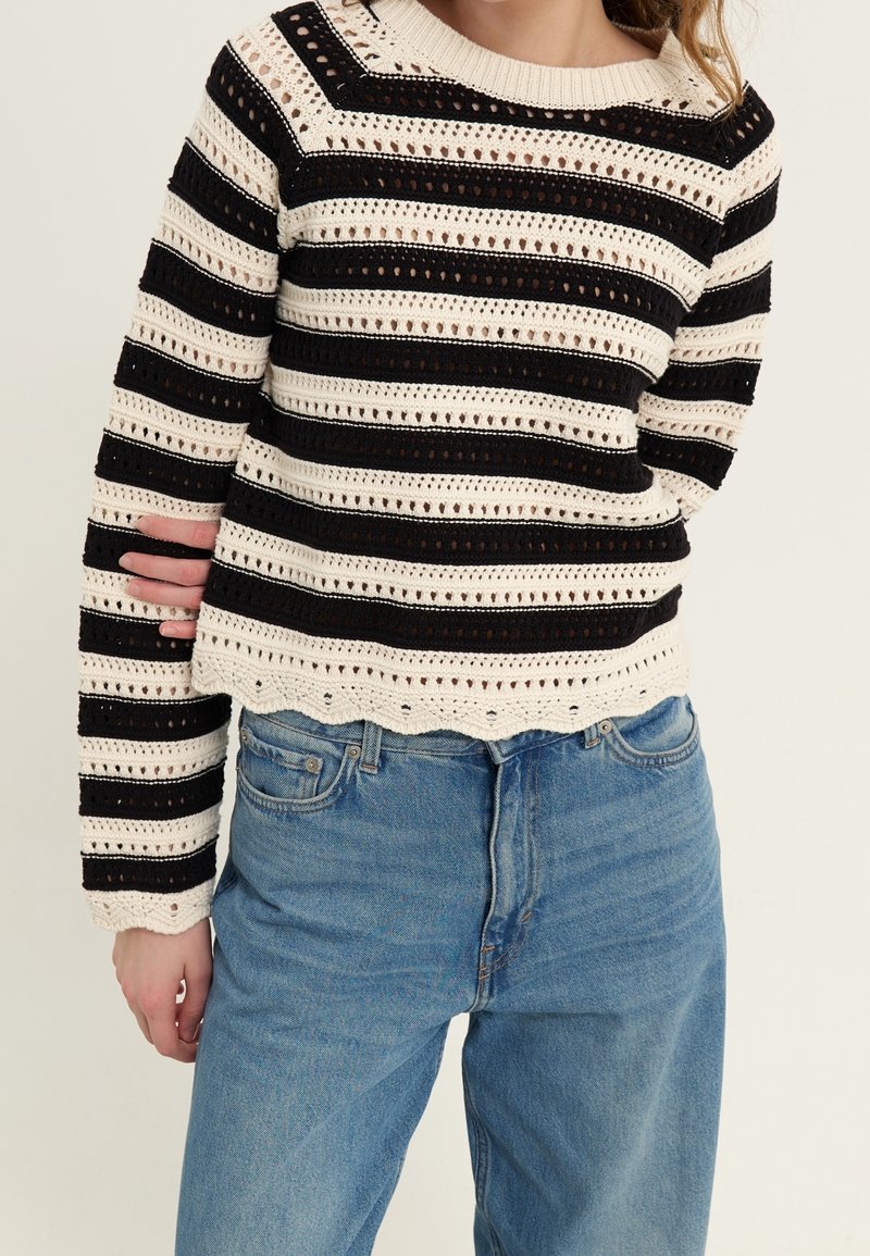Gehaakt sweater met zwarte en crèmekleurige strepen. Heeft een open brei ontwerp en een gegolfde zoom. Gecombineerd met lichtblauwe denim jeans.