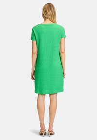 Grün kurzärmeliges Kleid mit strukturierter Oberfläche, knielanger Schnitt und rundem Ausschnitt. Trägt neutrale High-Heels.
