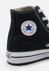 Svarte kanvas høye sneakers med hvit gummisåle, med en stjernelogo-lapp og hvite snøringer. Kontraststing på overdelen.