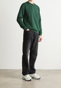 Felpa verde con scollo rotondo, abbinata a jeans neri a gamba dritta e sneakers bianche con accenti verdi e viola.