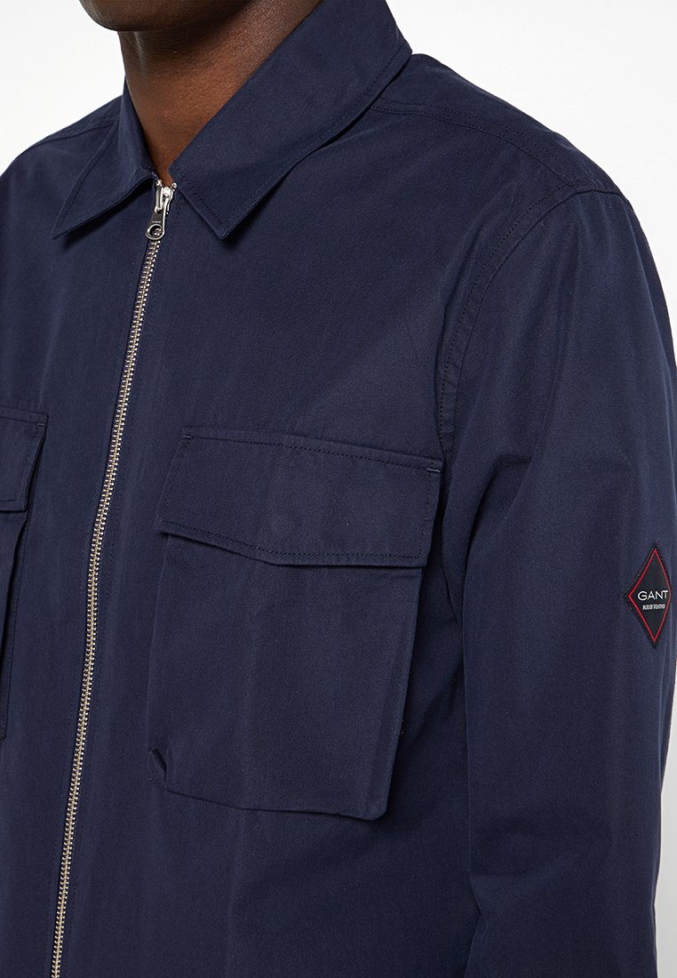 Hombre que lleva una chaqueta azul marino con cremallera, con grandes bolsillos de solapa y un parche con un logo en forma de diamante en la parte superior de la manga.