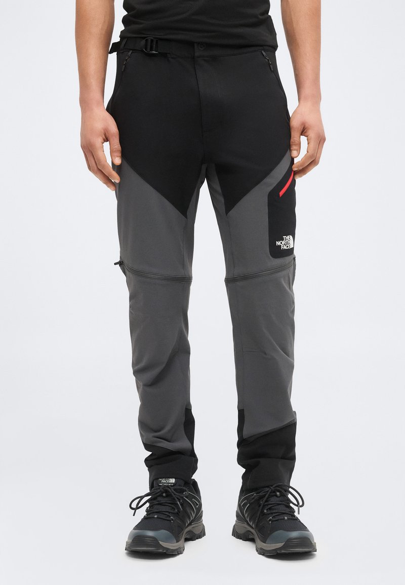 Mann trägt schwarze und graue North Face Zip-off Outdoorhose mit schwarzen Wanderschuhen, steht vor hellem Hintergrund.