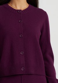 Cardigan lavorato a maglia viola con scollatura tonda, orlo a coste e sei bottoni abbinati sul davanti. Tessuto morbido con un fit accogliente.