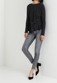 Top de manga larga gris oscuro con detalle de cintura anudada, combinado con jeans skinny grises brillantes y tacones negros de punta.