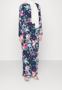 Kombinezon w kwiaty z długimi rękawami, granatowe tło, jasne, wielokolorowe kwiaty, elastyczna talia. Połączony z białą chustą i lekkimi sandałami.