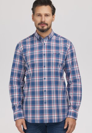 Mężczyzna o ciemnych włosach i brodzie, ubrany w niebiesko-biało-czerwoną kratowaną koszulę zapinaną na guziki, stojący na tle jednolitego jasnego tła.