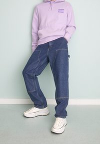 Lila hoodie med framficka och logotyp, kombinerad med lösa blå denimbyxor med sidofickor och kontrasterande sömmar, kompletterad med vita sneakers.