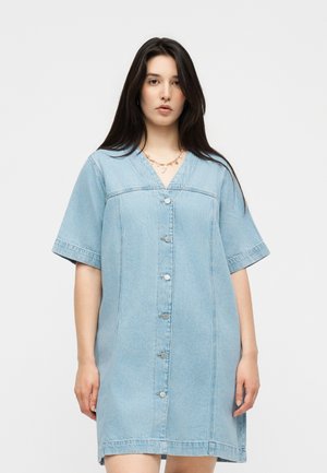 Kvinde med langt mørkt hår iført en lyseblå denim kjole med knapper, korte ærmer og et guldkæde.