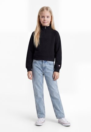 Ung pige med langt blondt hår iført en sort quarter-zip sweatshirt, lyseblå jeans og hvide sneakers, stående mod en hvid baggrund.