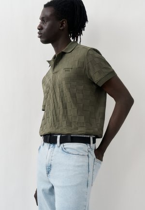 Νέος άνδρας με σκούρο δέρμα και dreadlocks, φορώντας μια ελαιόχρωμη πολο με υφή και ανοιχτό μπλε τζιν με μαύρη ζώνη, προφίλ από το πλάι.