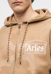 Άνδρας που φοράει μπεζ φούτερ με φερμουάρ με κορδόνια με μαύρες λεπτομέρειες και λευκό λογότυπο «Aries» πλαισιωμένο από το περίγραμμα κλασικού κτηρίου στο στήθος.