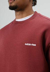 Sweat-shirt bordeaux à col rond, avec un logo brodé blanc "RAÍCES VIVAS" sur la poitrine. Texture de tissu lisse, coupe décontractée.
