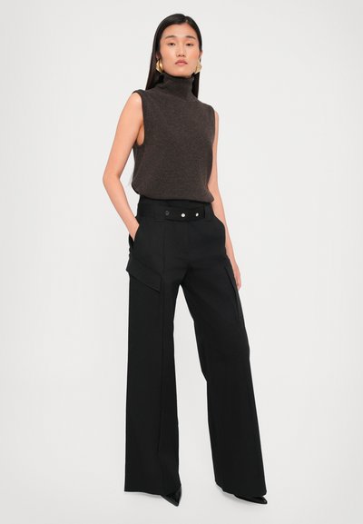 Brown sleeveless turtleneck sweater paired with high-waisted black wide-leg trousers featuring cargo pockets and a buttoned waist.