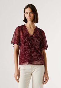 Blouse transparente bordeaux à pois blancs, col à volants, manches courtes évasées et détails de boutons sur le devant.