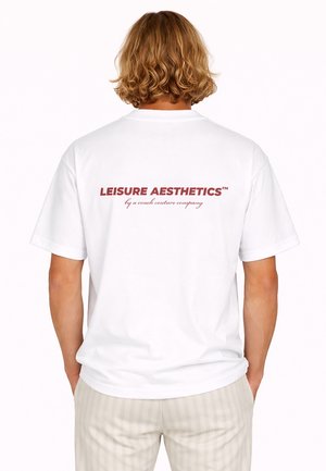 Person mit schulterlangem lockigem Haar, trägt ein weißes T-Shirt mit rotem Text "LEISURE AESTHETICS by a couch couture company" auf dem Rücken.
