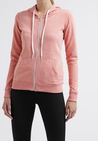 Mujer vestida con una sudadera con cremallera de color rosa claro, con cordones blancos y pantalones negros, de pie contra un fondo blanco liso.