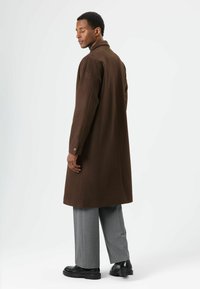 Bruine lange jas met een kraag, voorzien van verlaagde schouders en met knopen afsluitbare manchetten. Gecombineerd met grijze, wijde broekspijpen en zwarte schoenen.